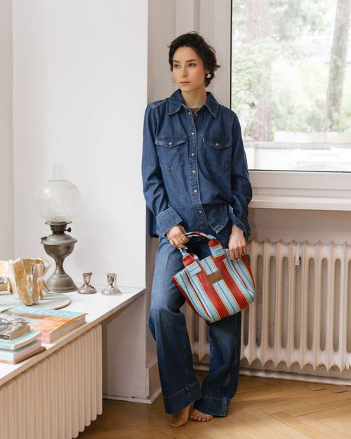 Woman in denim outfit holding a pink striped bag in a room with a window and radiator.
