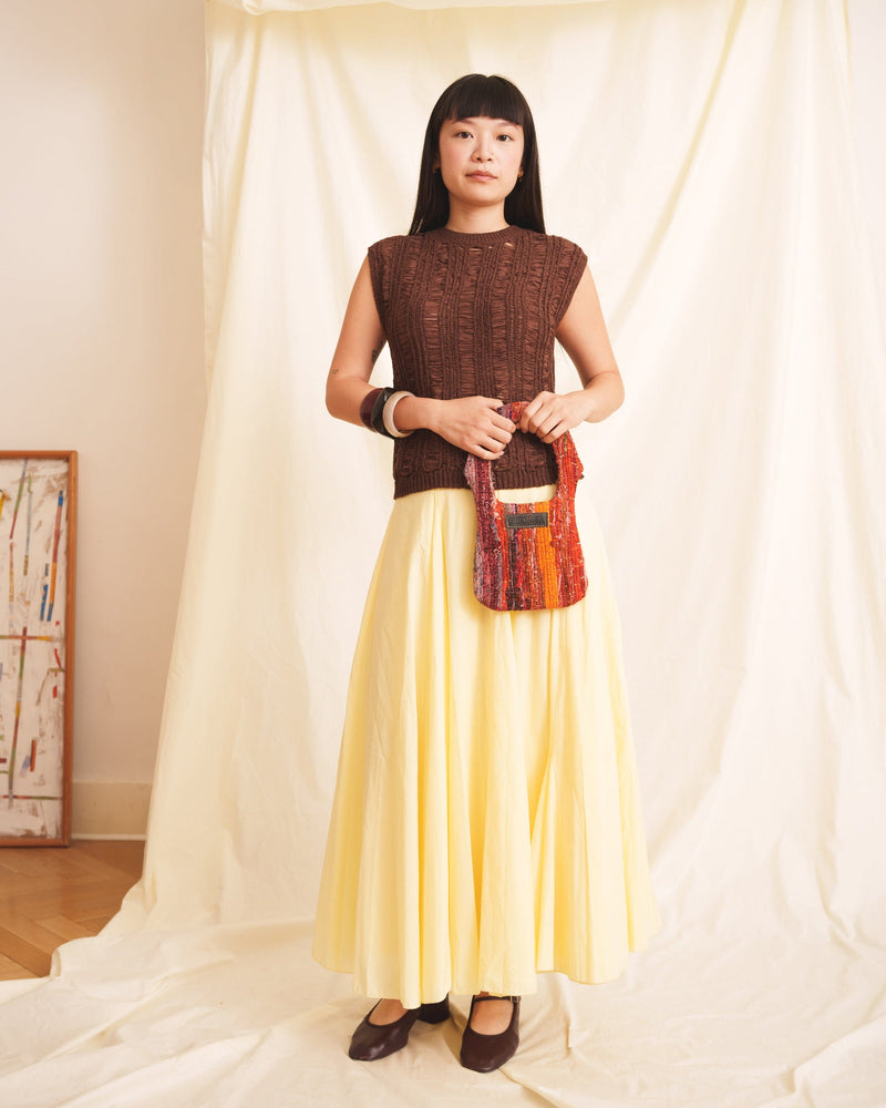 Woman wearing a brown top and yellow skirt holding a cute red shaded purse -handbag in her hand standing in front of white curtains.