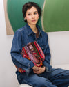 Person wearing a denim jacket holding a colorful bag against a green abstract painting.