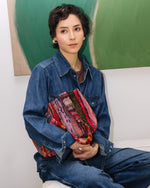 Person wearing a denim jacket holding a colorful bag against a green abstract painting.