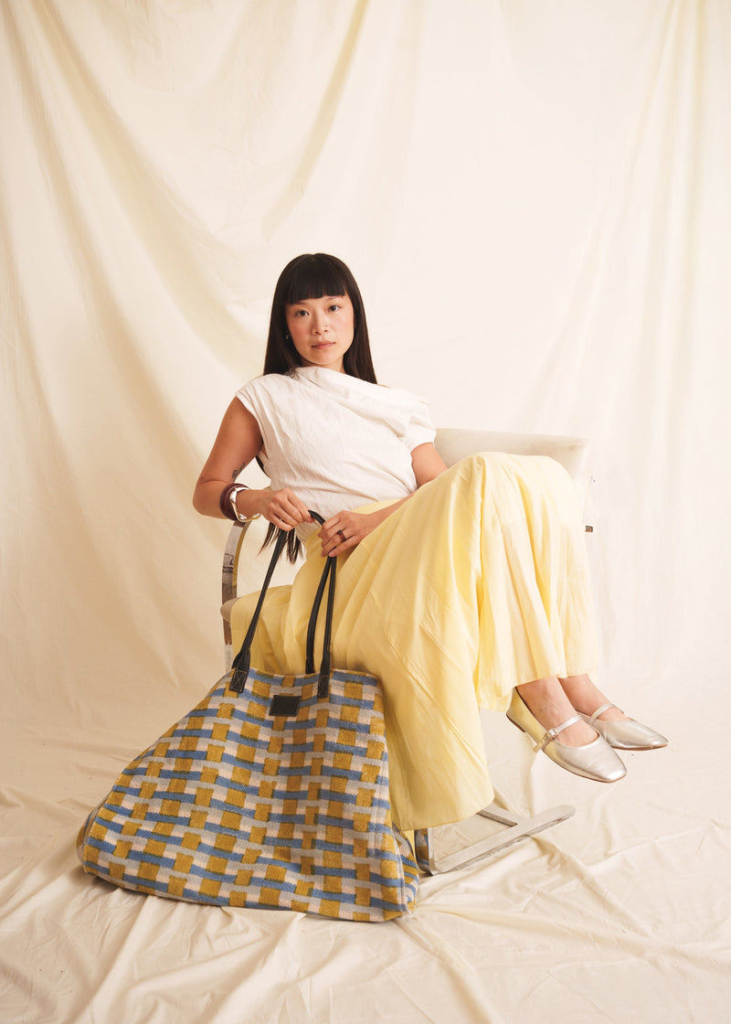 Woman sitting on a chair wearing a white top and yellow skirt, holding a blue checkered bag.